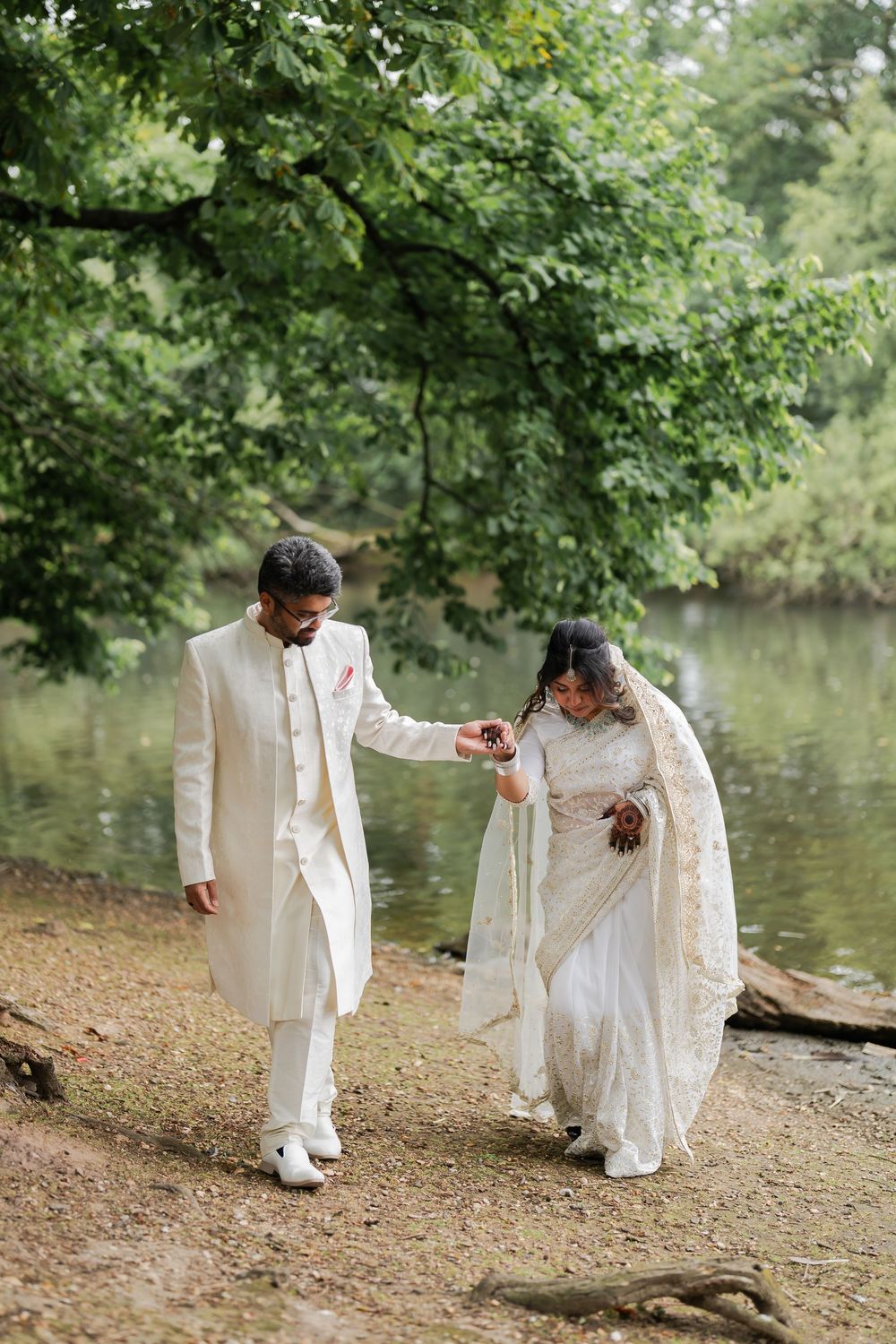 South Asian bride and groom in traditional white attire walk hand in hand along a serene lakeside path surrounded by lush greenery.