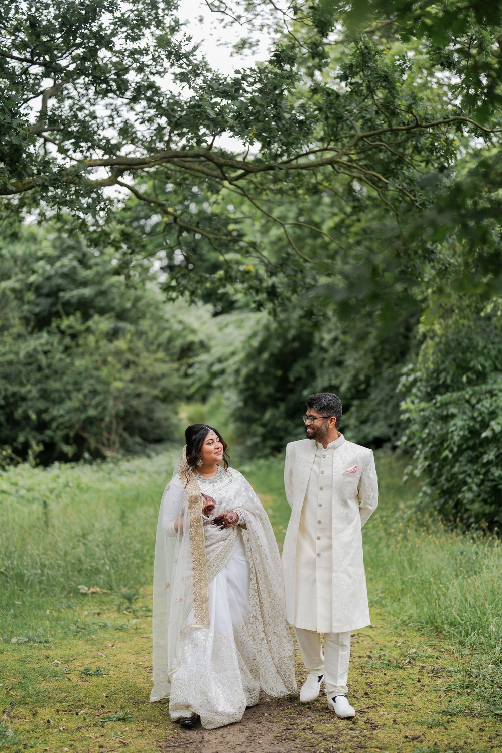 A couple in traditional South Asian wedding attire walks through a lush green garden path, smiling at each other.
