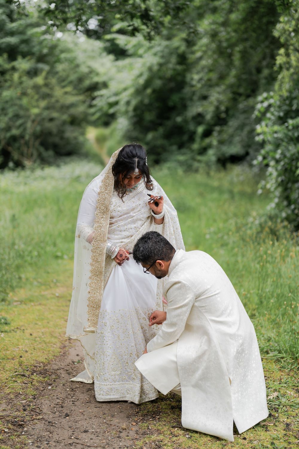 Groom in white sherwani kneels to fix bride's white embroidered lehenga on a garden path.