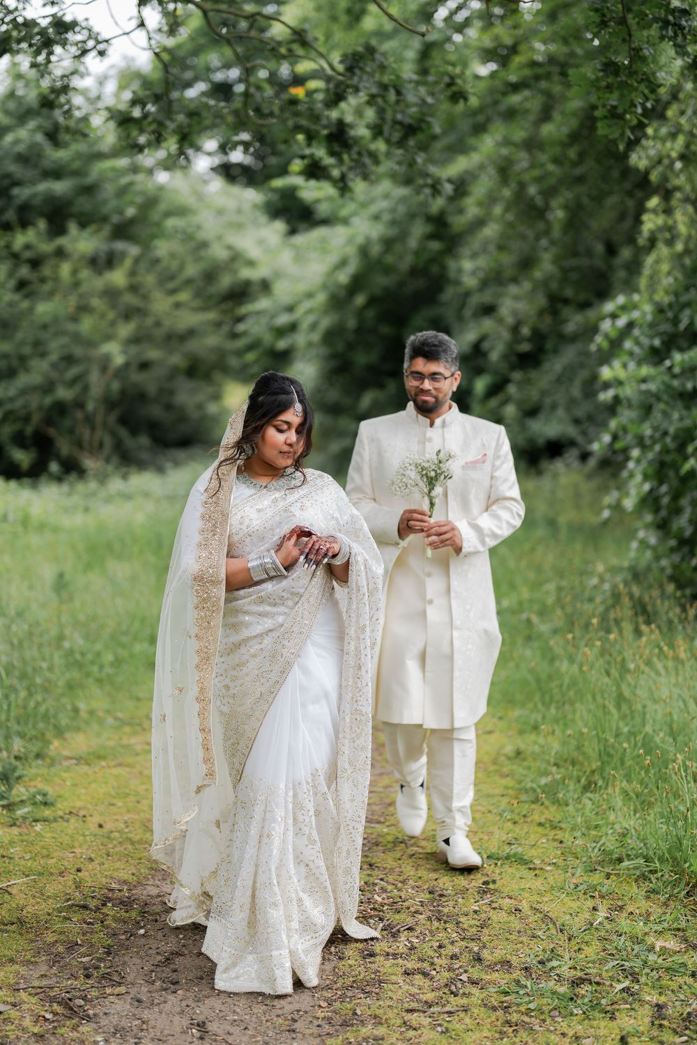South Asian couple in white wedding attire walk a garden path, bride in embroidered saree, groom in sherwani holding flowers.