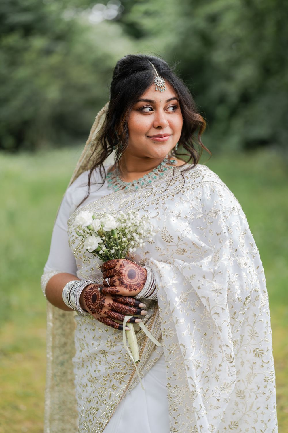 Indian bride in white saree with mehndi hands holding white bouquet outdoors.