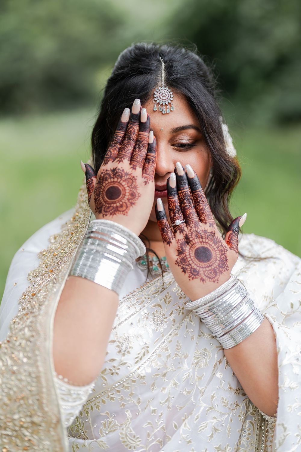 Indian bride in white saree displaying intricate henna mehndi designs on hands, wearing silver bangles and maang tikka.