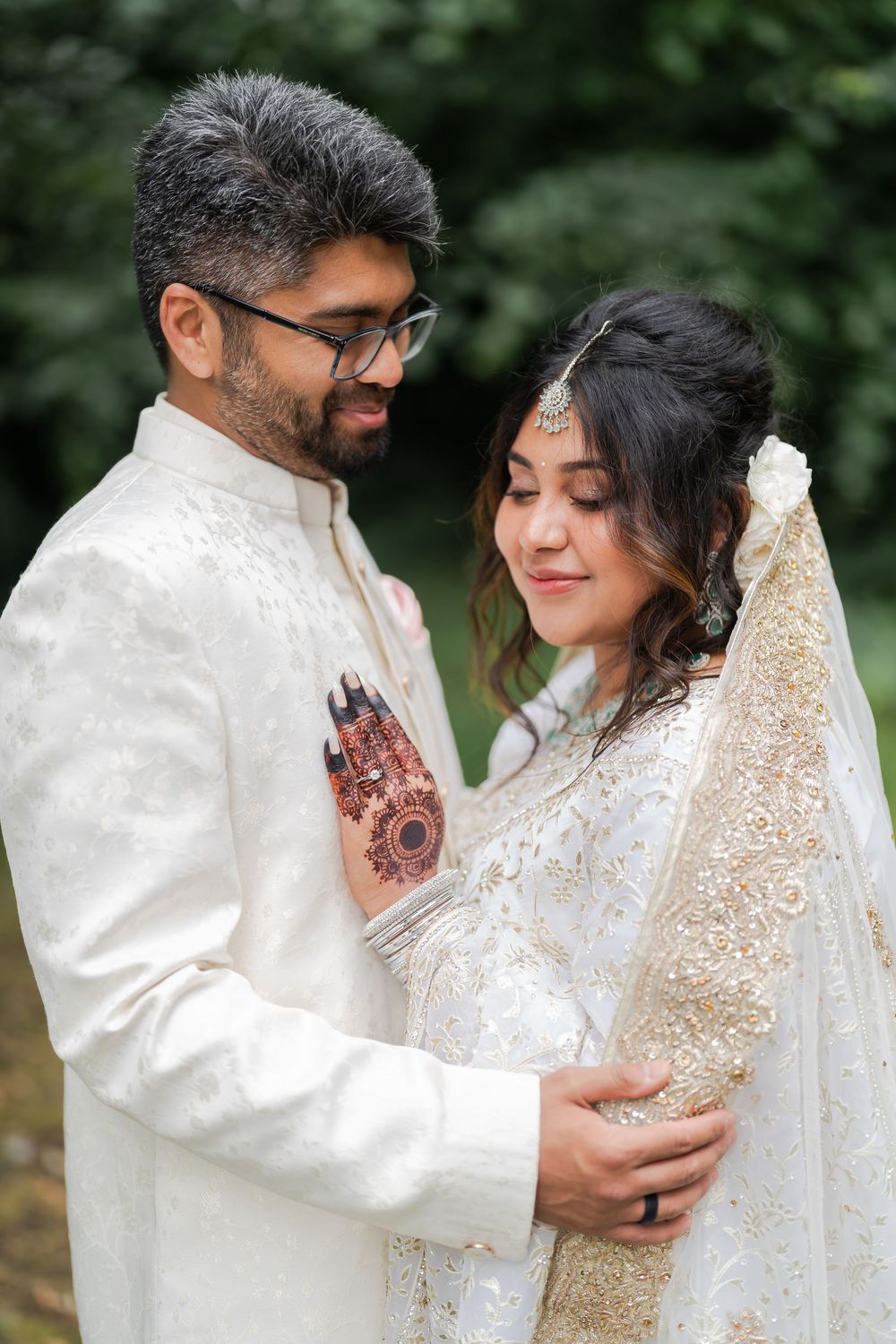 South Asian couple in white wedding attire embrace outdoors, bride wearing mehndi and veil, groom in sherwani with glasses.