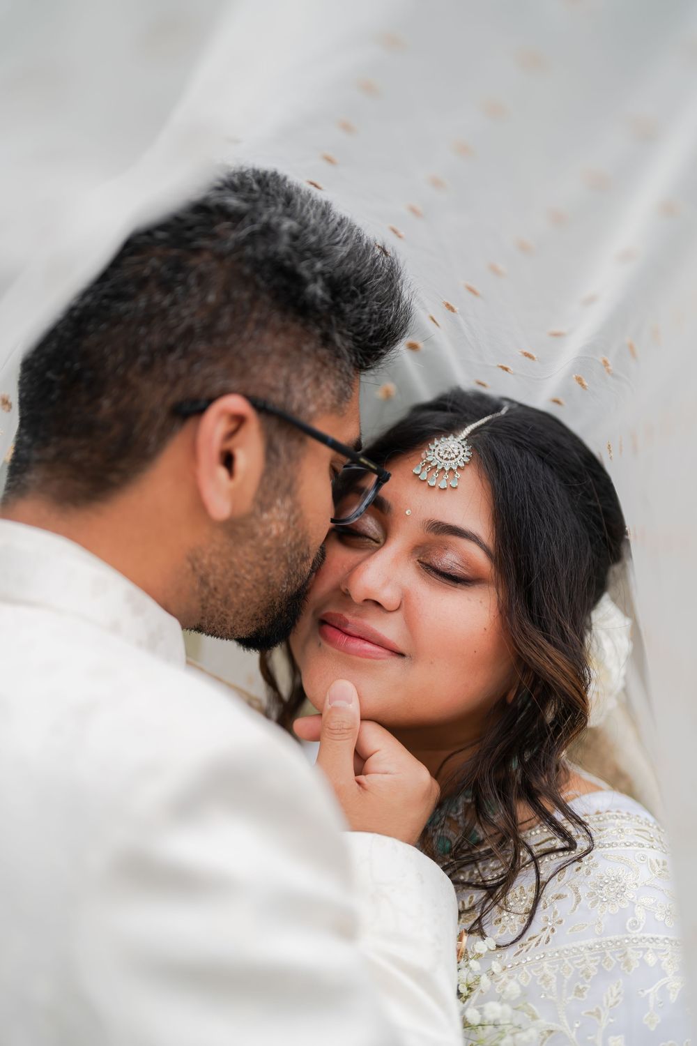 South Asian bride and groom share an intimate moment under a white veil, bride wearing traditional maang tikka jewelry.
