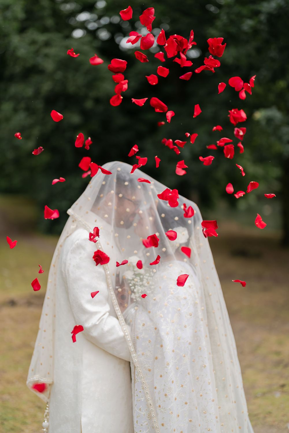 Bride and groom in white attire embracing under a shower of red rose petals outdoors.