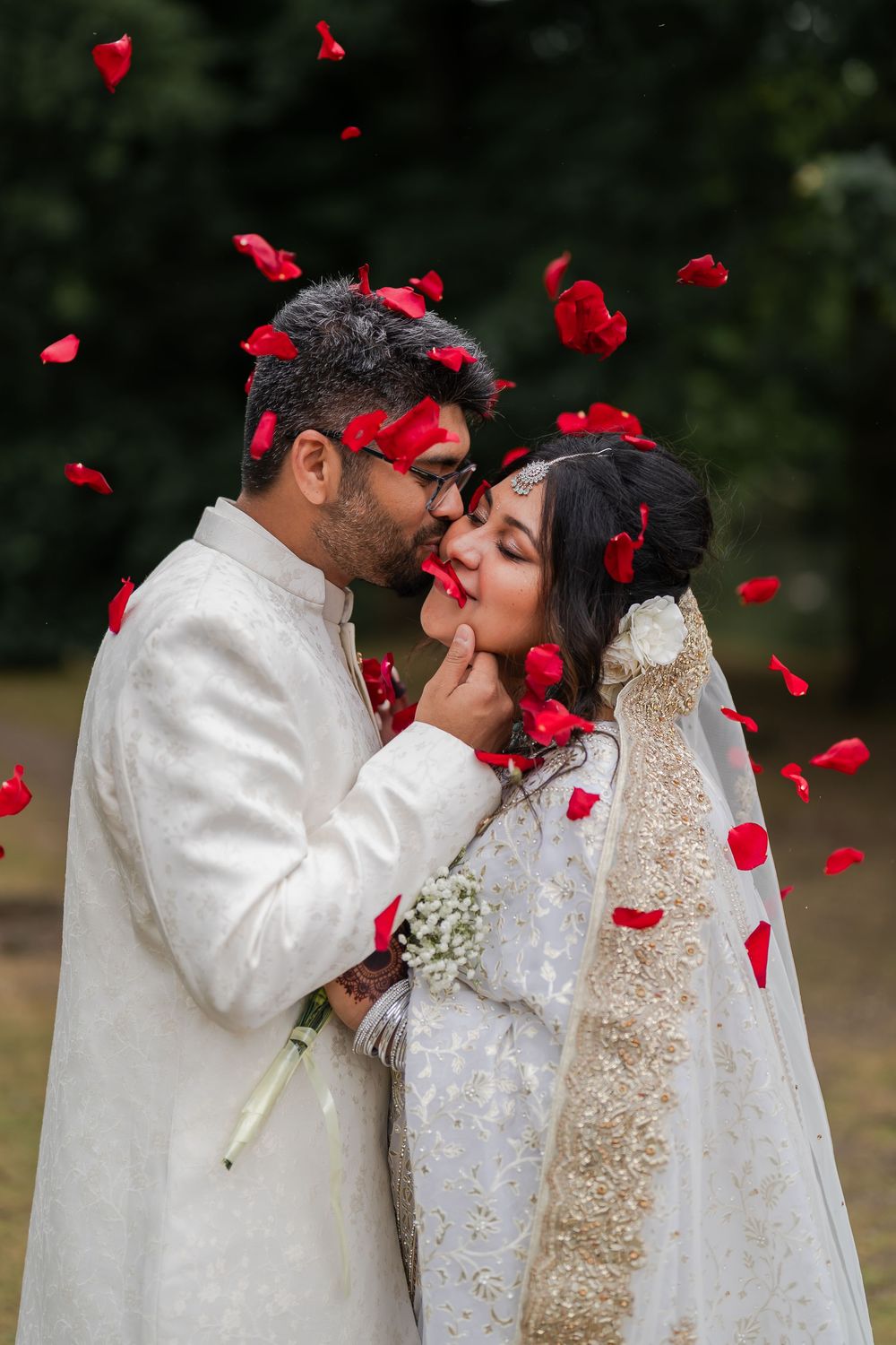 South Asian wedding couple kissing as red rose petals fall around them, both dressed in elegant white traditional attire.