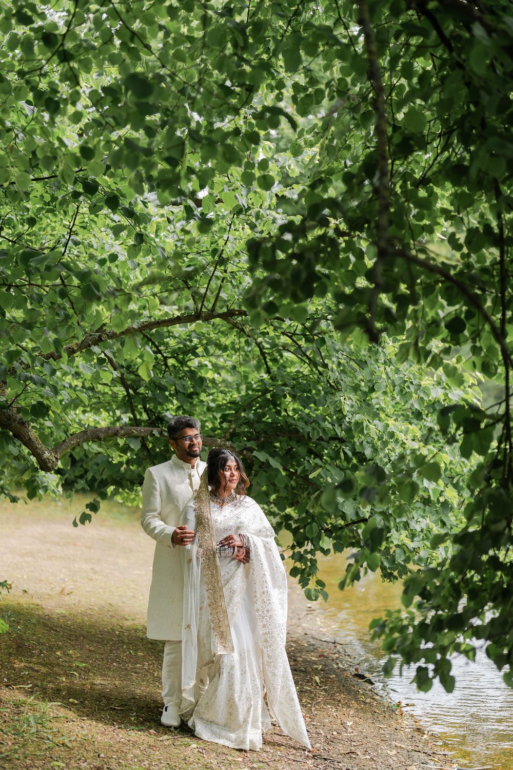 South Asian bride and groom in white traditional attire walking along a scenic lakeside path beneath lush green trees.