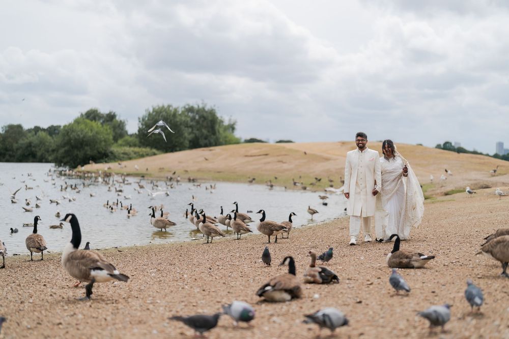 Couple in white traditional attire walking along a sandy lakeside shore surrounded by geese and pigeons under cloudy skies.