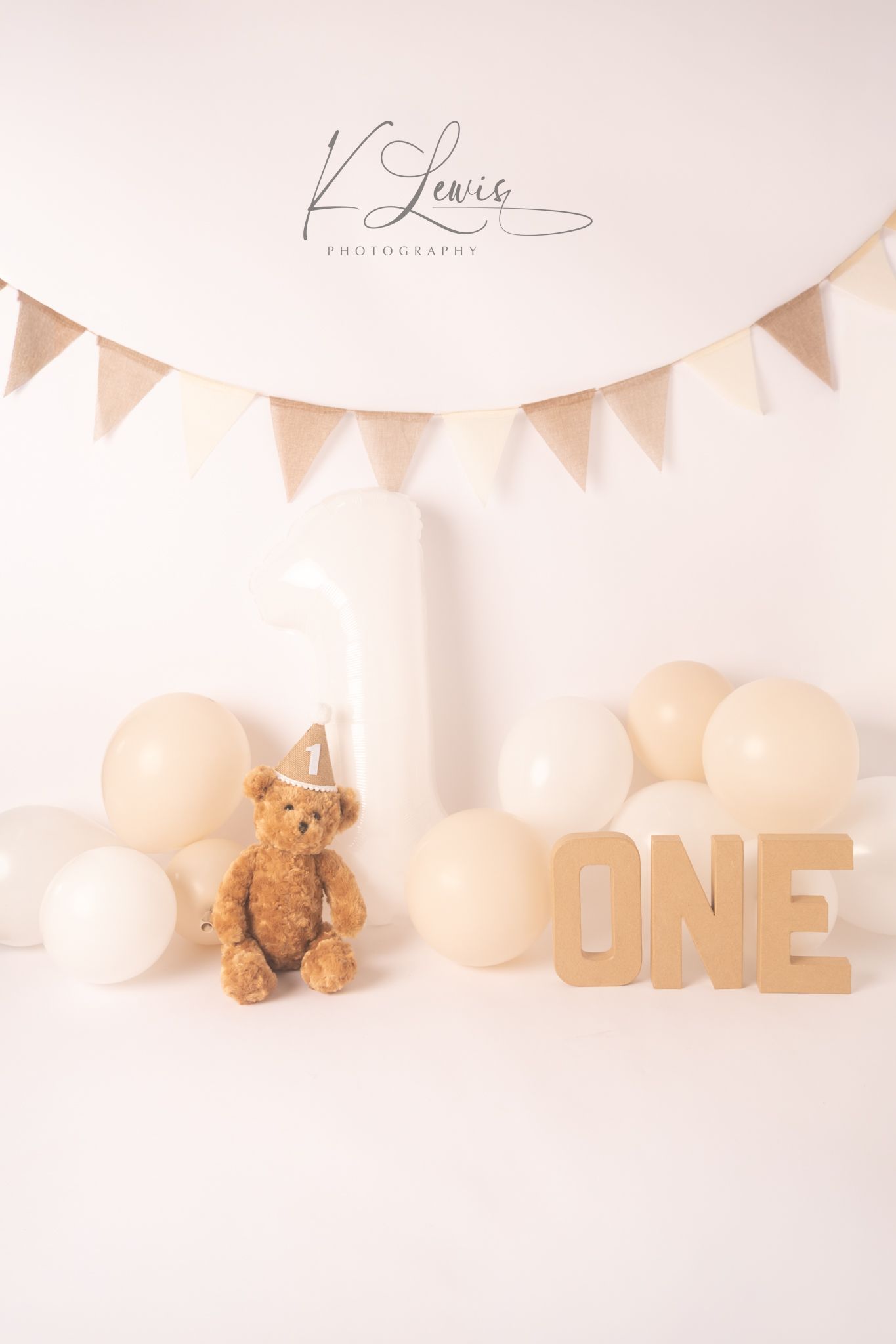 A first birthday cake smash setup with a teddy bear, white number one, wooden ONE letters, cream balloons, and burlap bunting.