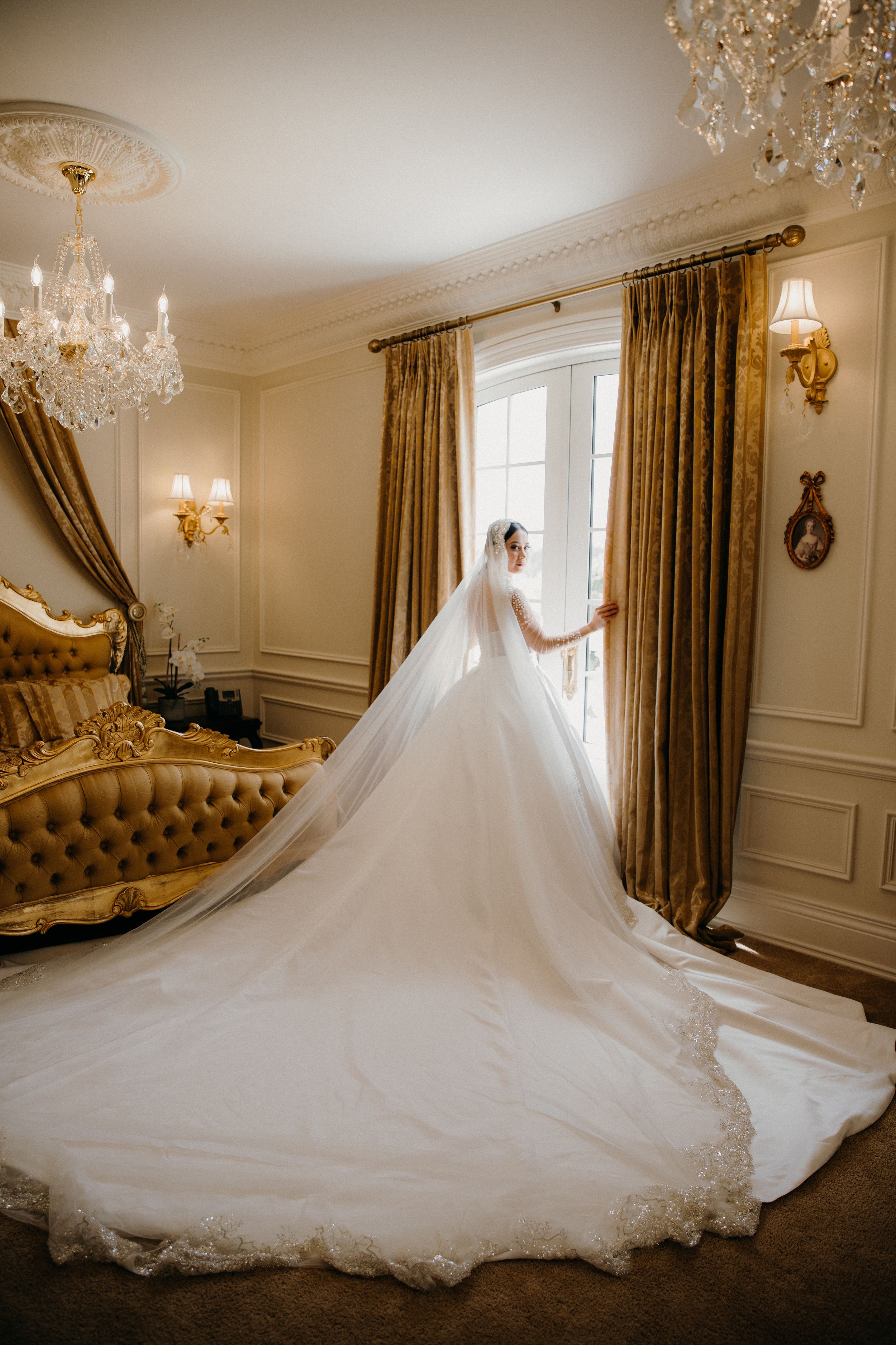 Elegant wedding dress with dramatic cathedral train flows across luxury bridal suite with crystal chandelier and gold decor.