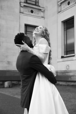 Black and white photo of newlyweds sharing joyful embrace outside classic building.