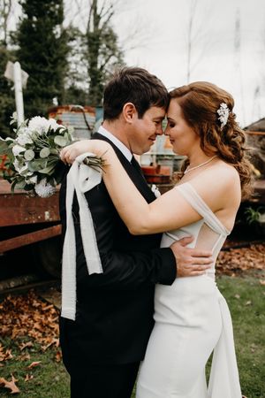 Romantic wedding couple embraces outdoors with bride holding white bouquet against rustic wooden backdrop.