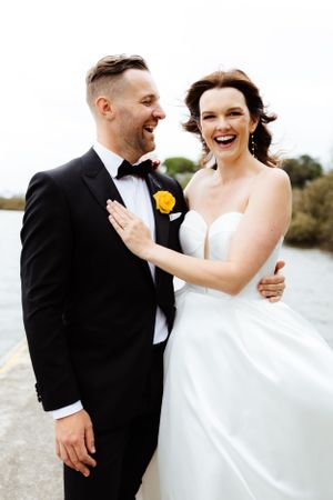 A joyful couple in wedding attire laugh together outdoors by the water with the bride in white and groom in black tuxedo.