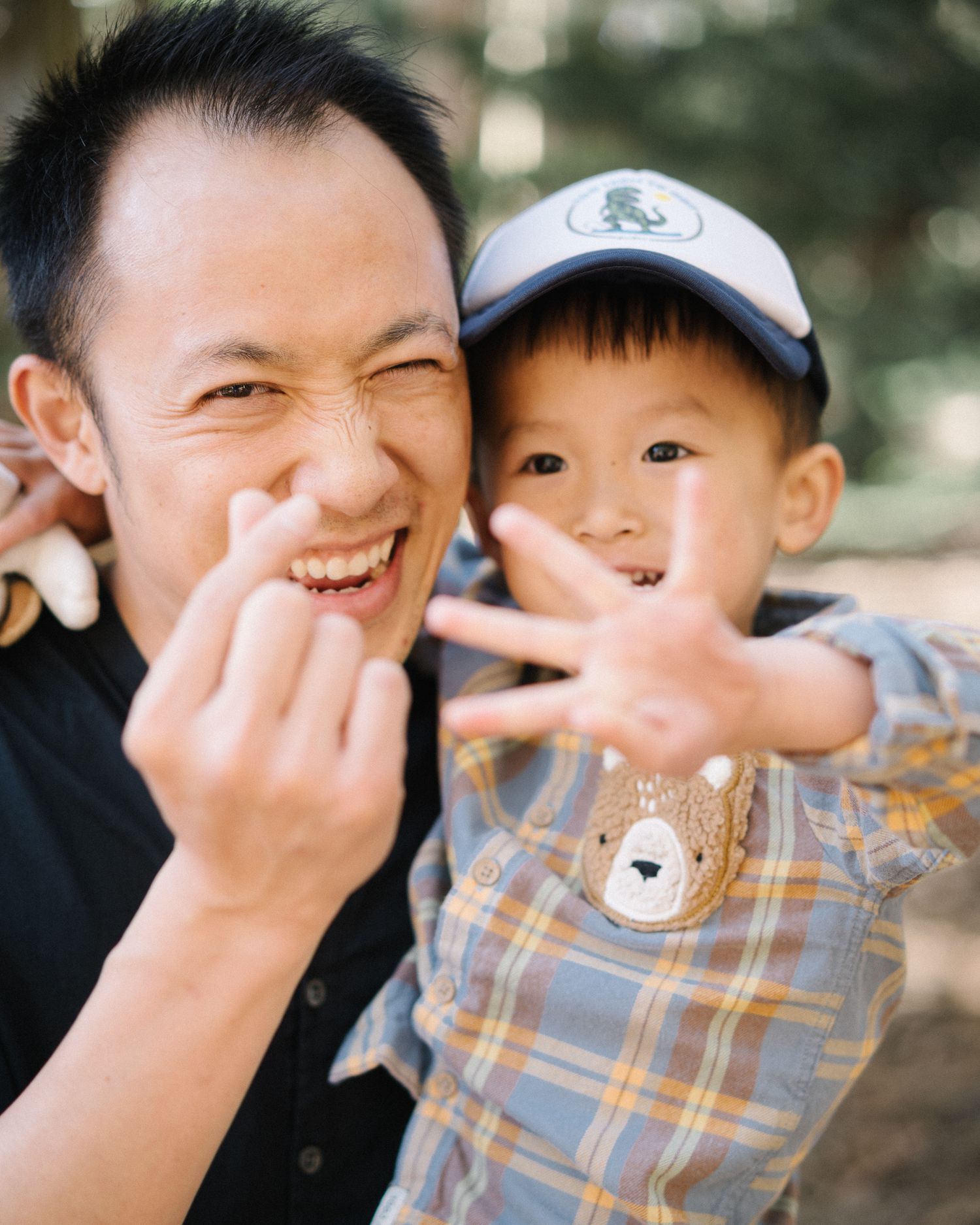 A man and young boy in a plaid shirt and bear cap smile and gesture playfully at the camera outdoors.