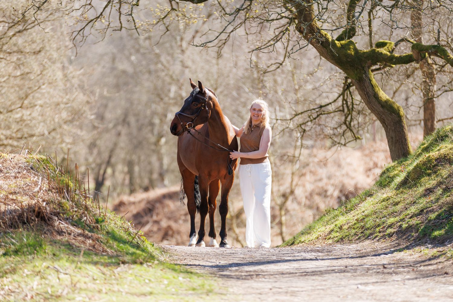 Young girl standing between a chestnut and grey horse near ornate stone gates in a lush green estate.