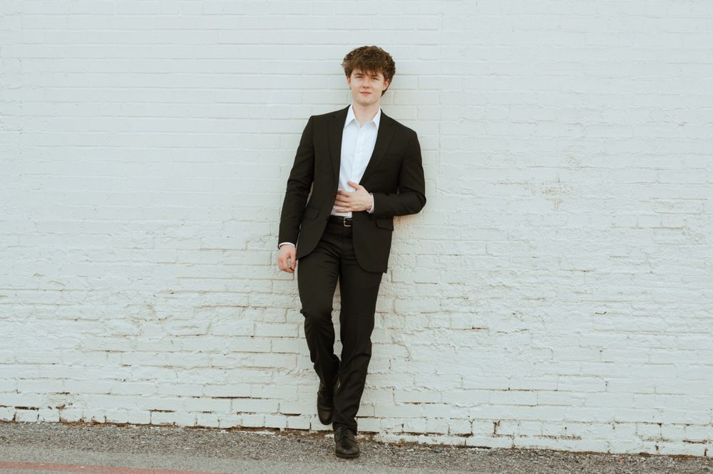 Young man in black suit leaning against white brick wall in professional portrait photo.