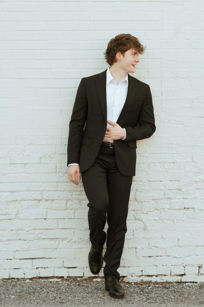 Young man in dark suit and white shirt smiling, leaning against white brick wall in a professional portrait.