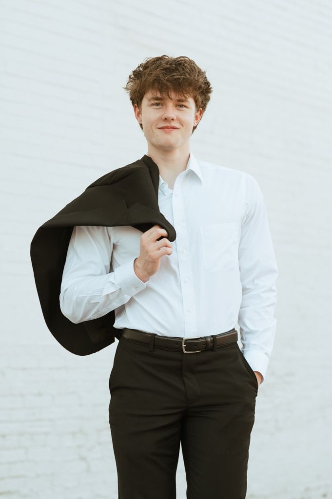 Young man in white dress shirt holding dark blazer over shoulder, smiling confidently against white brick wall.