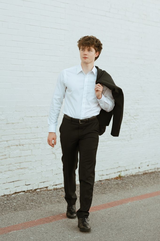 Young man in white dress shirt carrying black blazer over shoulder walks confidently along brick wall.