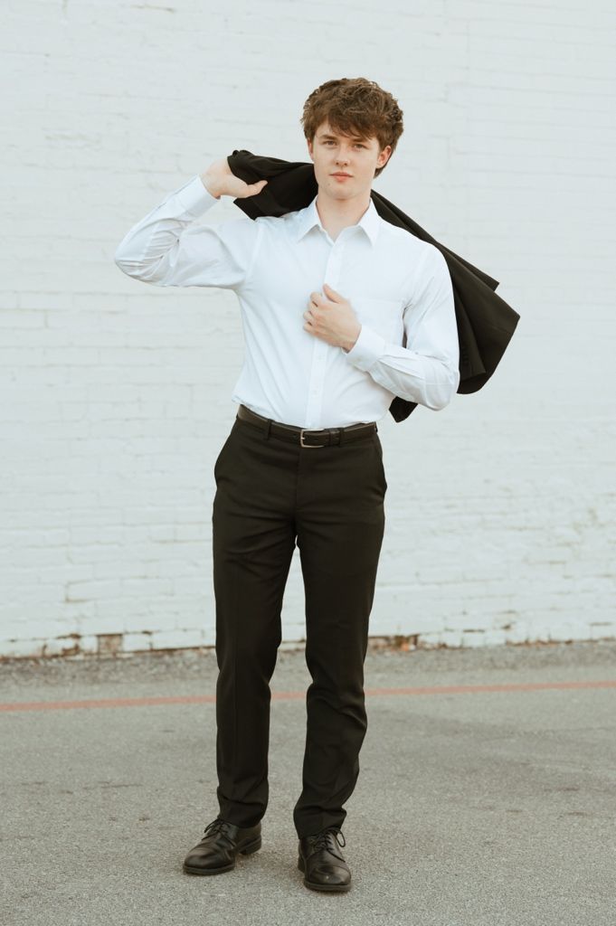 Young man in white dress shirt and black slacks draping suit jacket over shoulder against white brick wall.