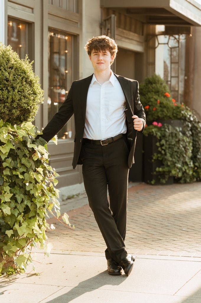 Young man in black suit poses confidently on sunny sidewalk beside lush green bushes outside elegant building.