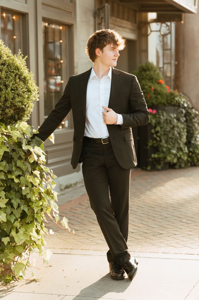 Young man in black suit walking outdoors holding jacket lapel near ivy-covered wall.