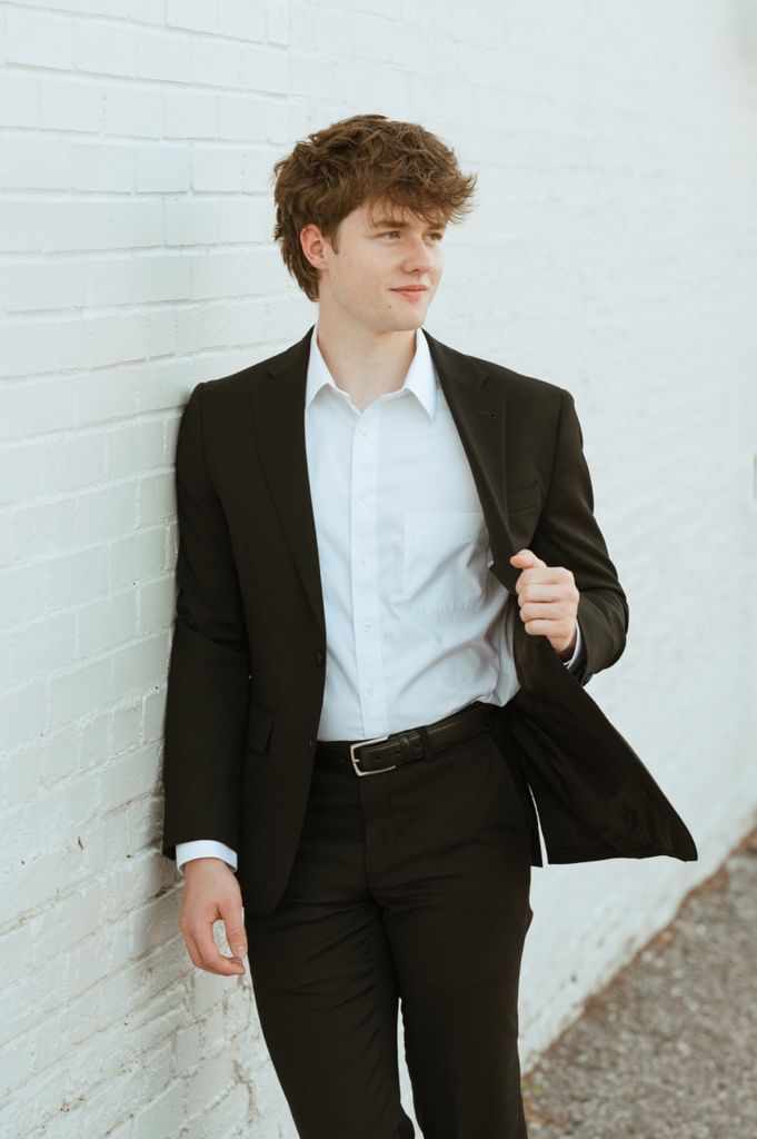 Young man in black suit leaning against white brick wall, holding lapel confidently.