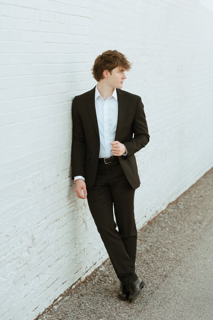 Young man in dark suit and white dress shirt leaning against white brick wall outdoors.