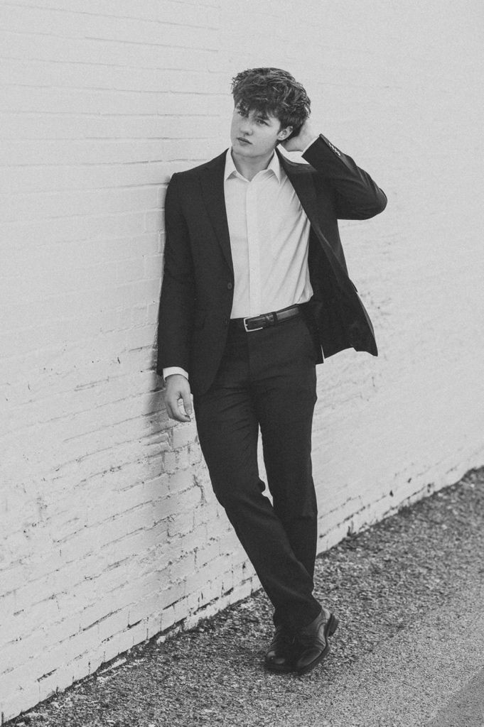 Young man in black suit leaning against white brick wall, black and white portrait photo.