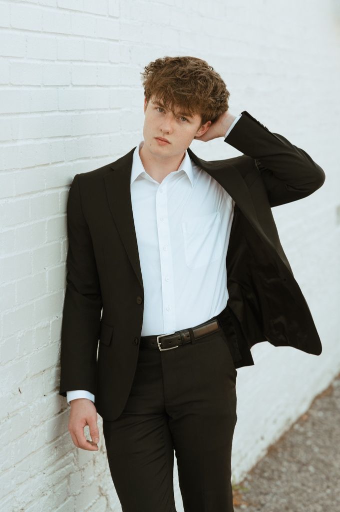 Young man in black suit and white shirt leaning against white brick wall with hand behind head.