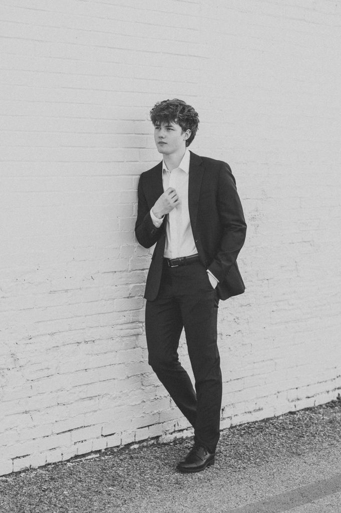 Young man in black suit leaning against white brick wall in black and white portrait photo.