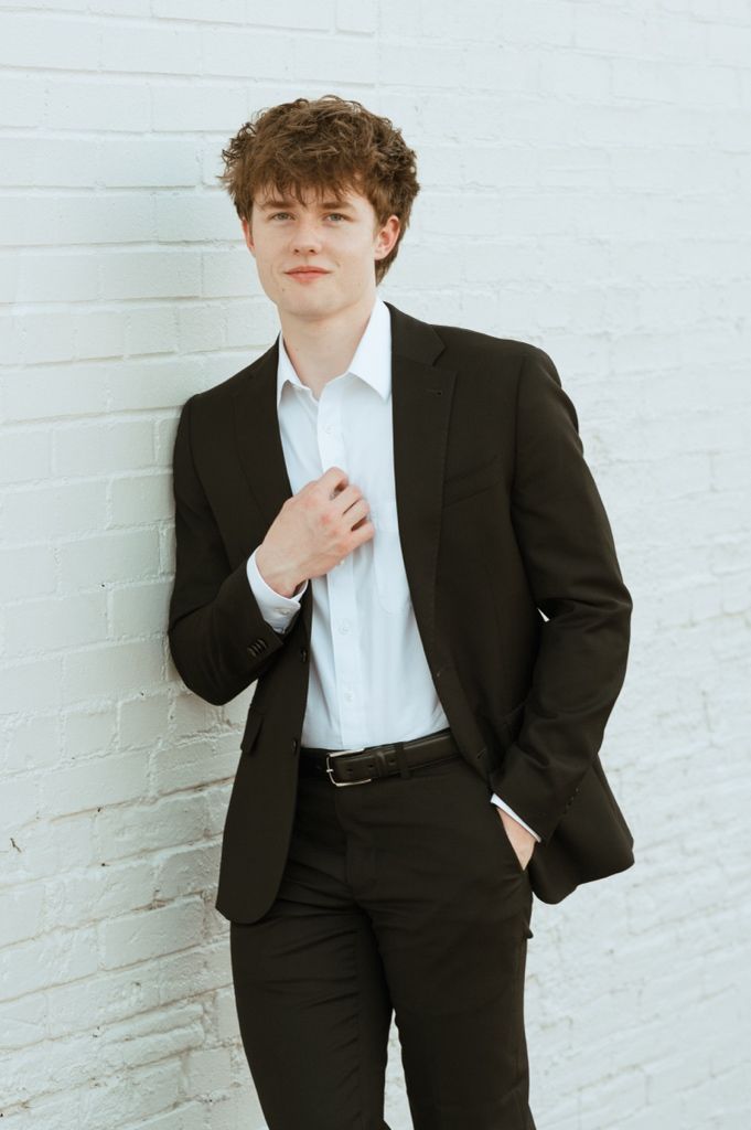 Young man in black suit and white shirt leaning against white brick wall, posing confidently with hand in pocket.