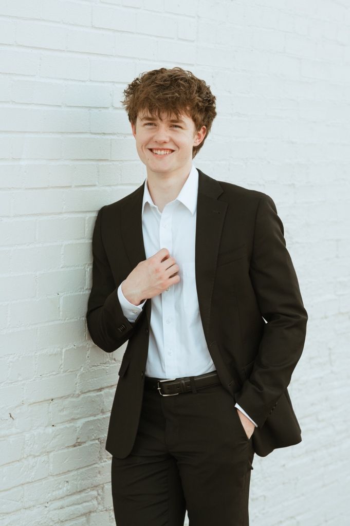 Young man in black suit and white shirt smiling against white brick wall.