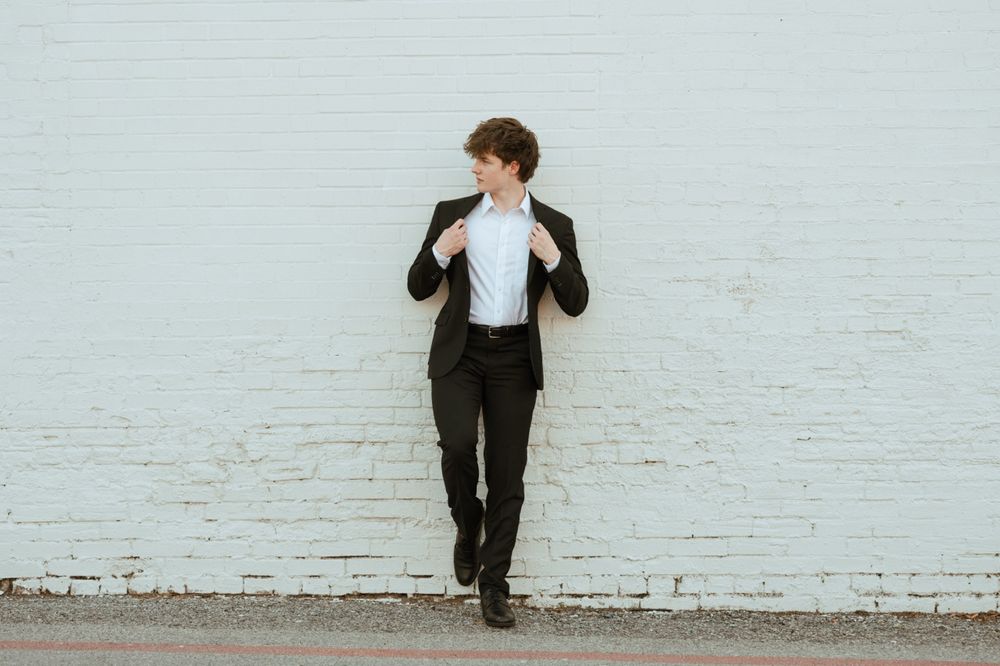 Young man in black suit leaning against white brick wall, adjusting jacket collar outdoors.