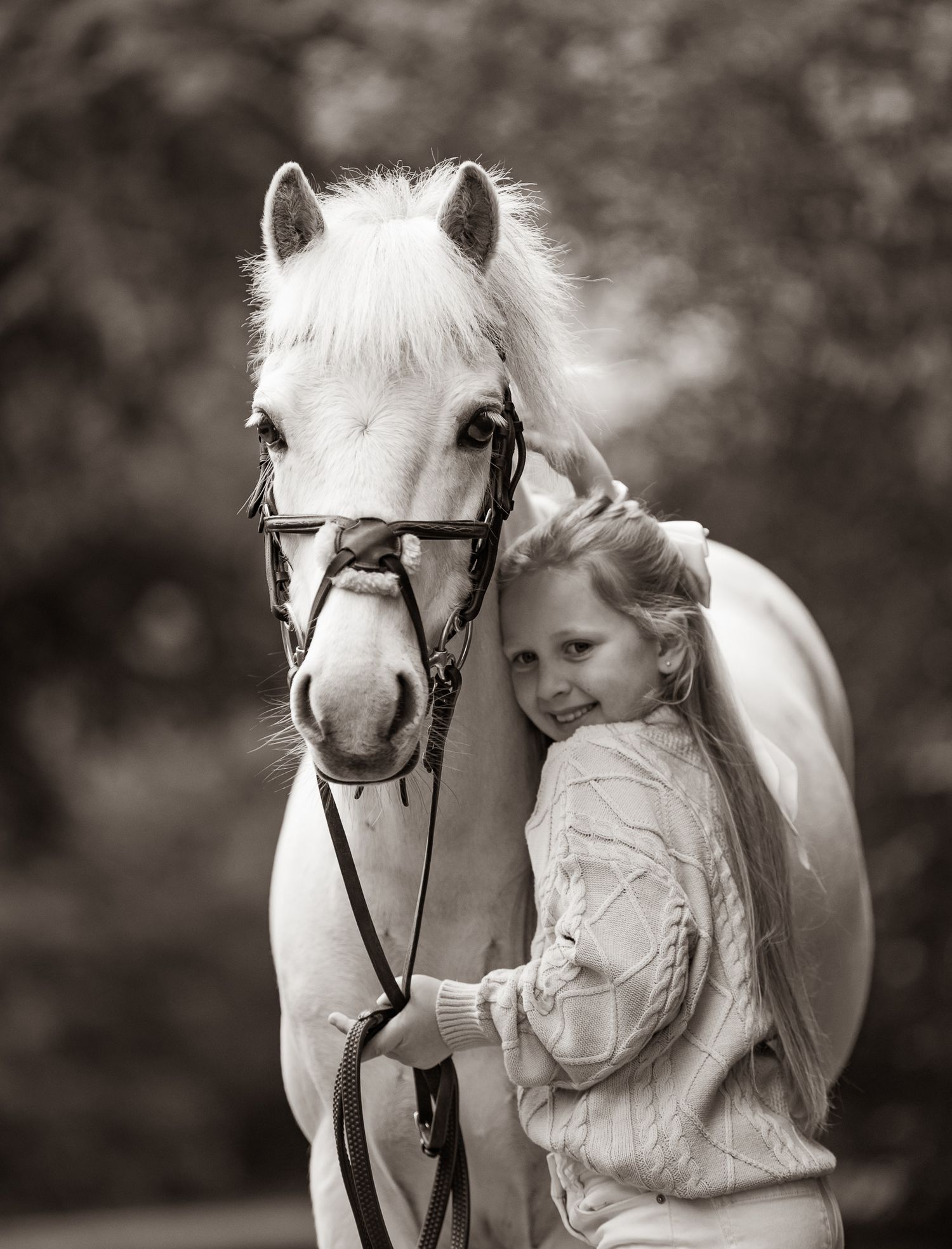 A young girl smiles warmly while hugging a white pony wearing a bridle in this charming black and white portrait.