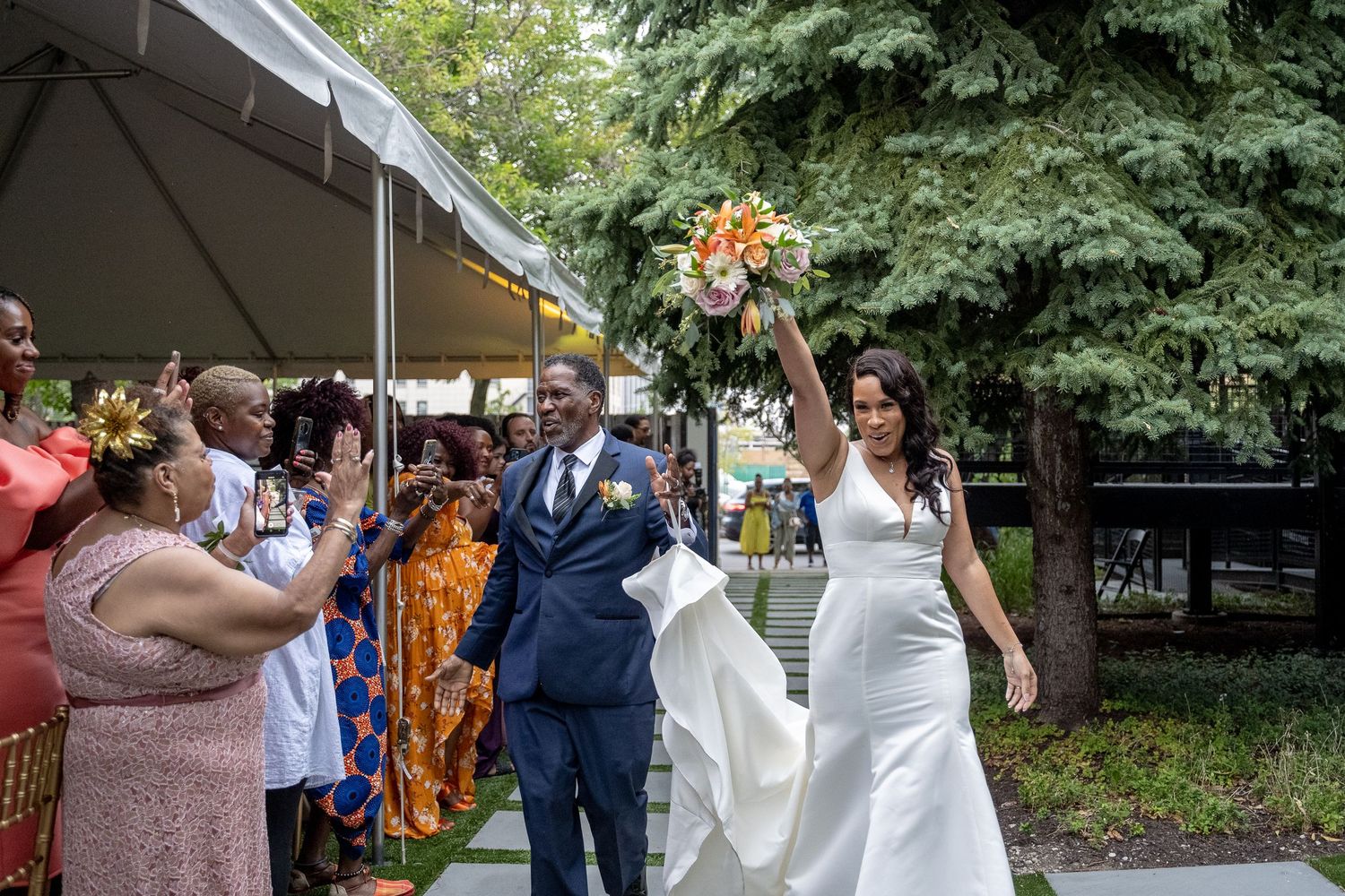 Bride raises bouquet of orange flowers while walking with groom past guests in colorful attire at outdoor wedding.