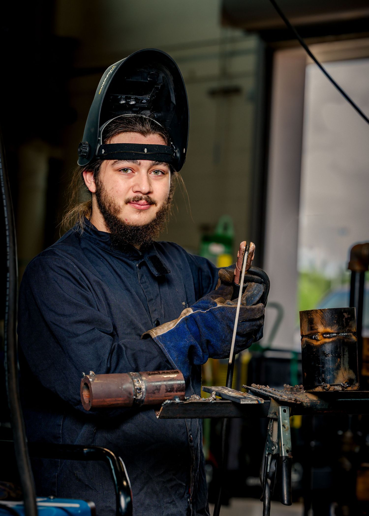 Young male welder with beard wearing protective mask and blue work uniform holding welding tools in workshop.