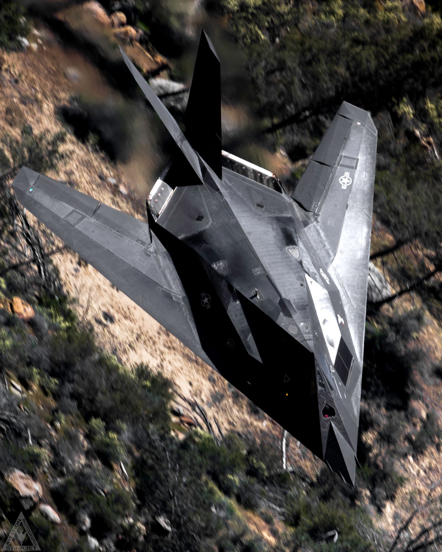 An F-117 Knighthawk flies low level against a background of mountain shrubbery and dirt