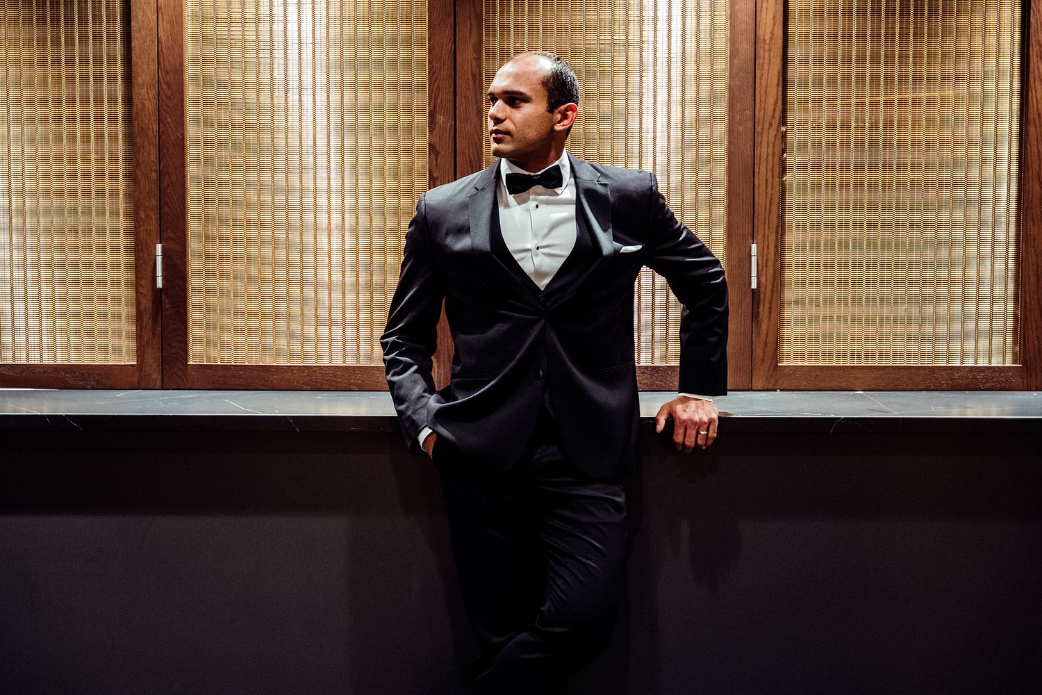 Elegant man in a black tuxedo with bow tie leaning against a wall in a sophisticated indoor setting.