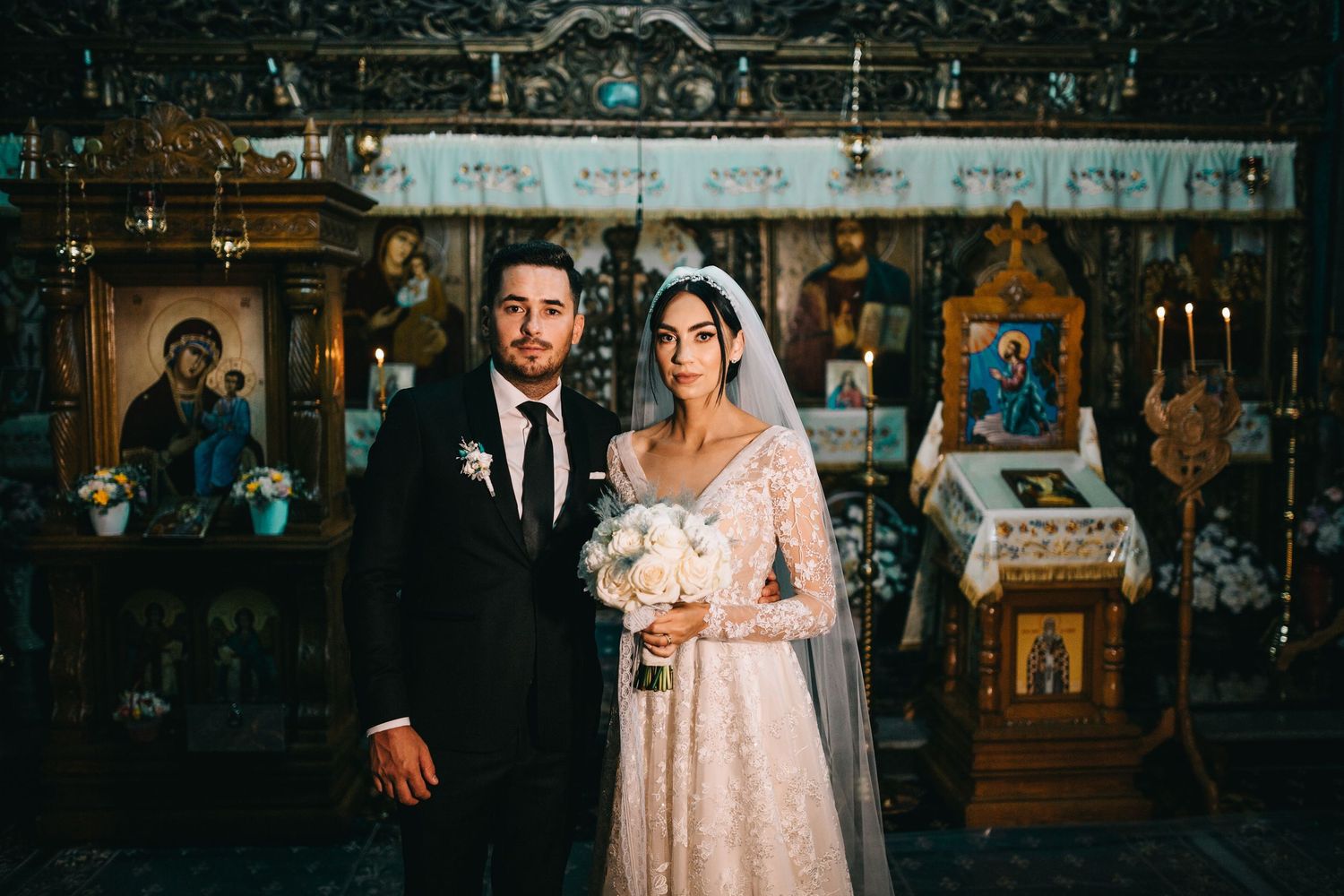 Bride and groom share kiss inside ornate Orthodox church beneath crystal chandelier and golden icons.