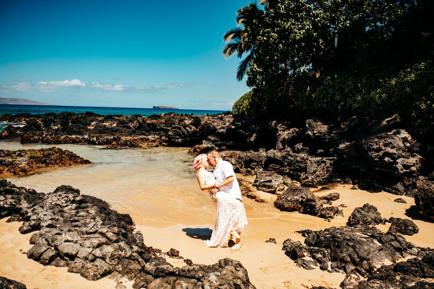 Couple embracing on a sandy beach surrounded by black lava rocks and turquoise ocean in Hawaii.