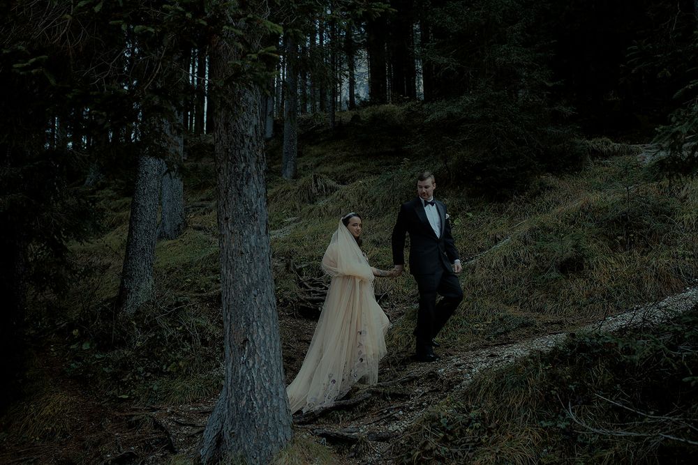 Bride in flowing gown and groom in tuxedo walk through a dark, moody forest during their wedding elopement.