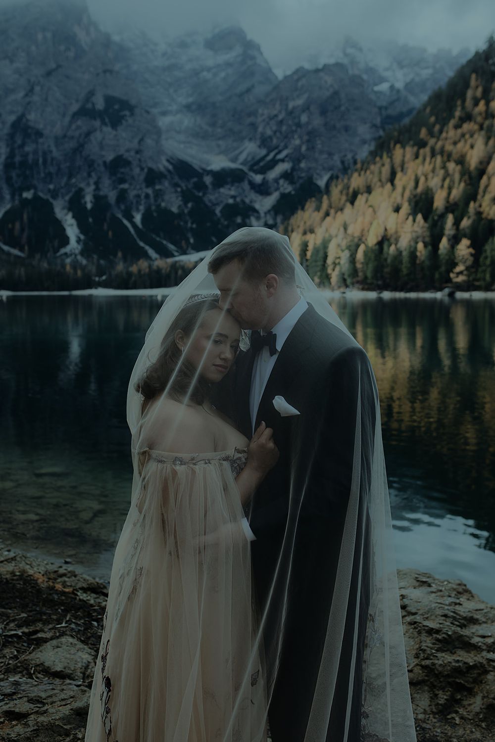 A bride and groom share an intimate moment under a veil by a serene alpine lake with autumn trees and mountains.