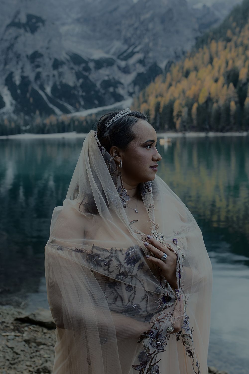 Bride in floral gown and veil stands by a mountain lake with autumn trees and rocky peaks in background.