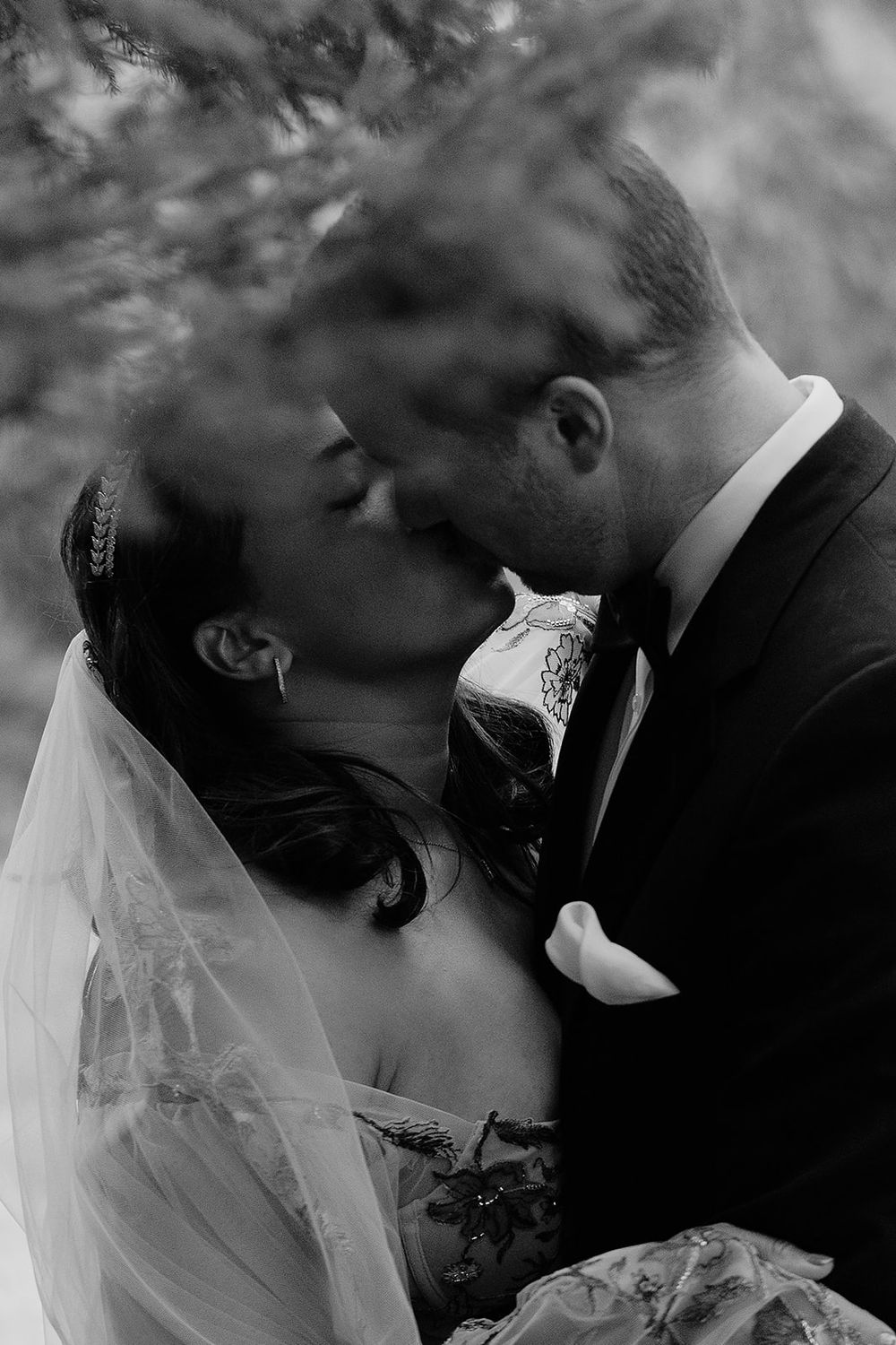 Black and white photo of a bride and groom sharing a romantic kiss under lush trees on their wedding day.