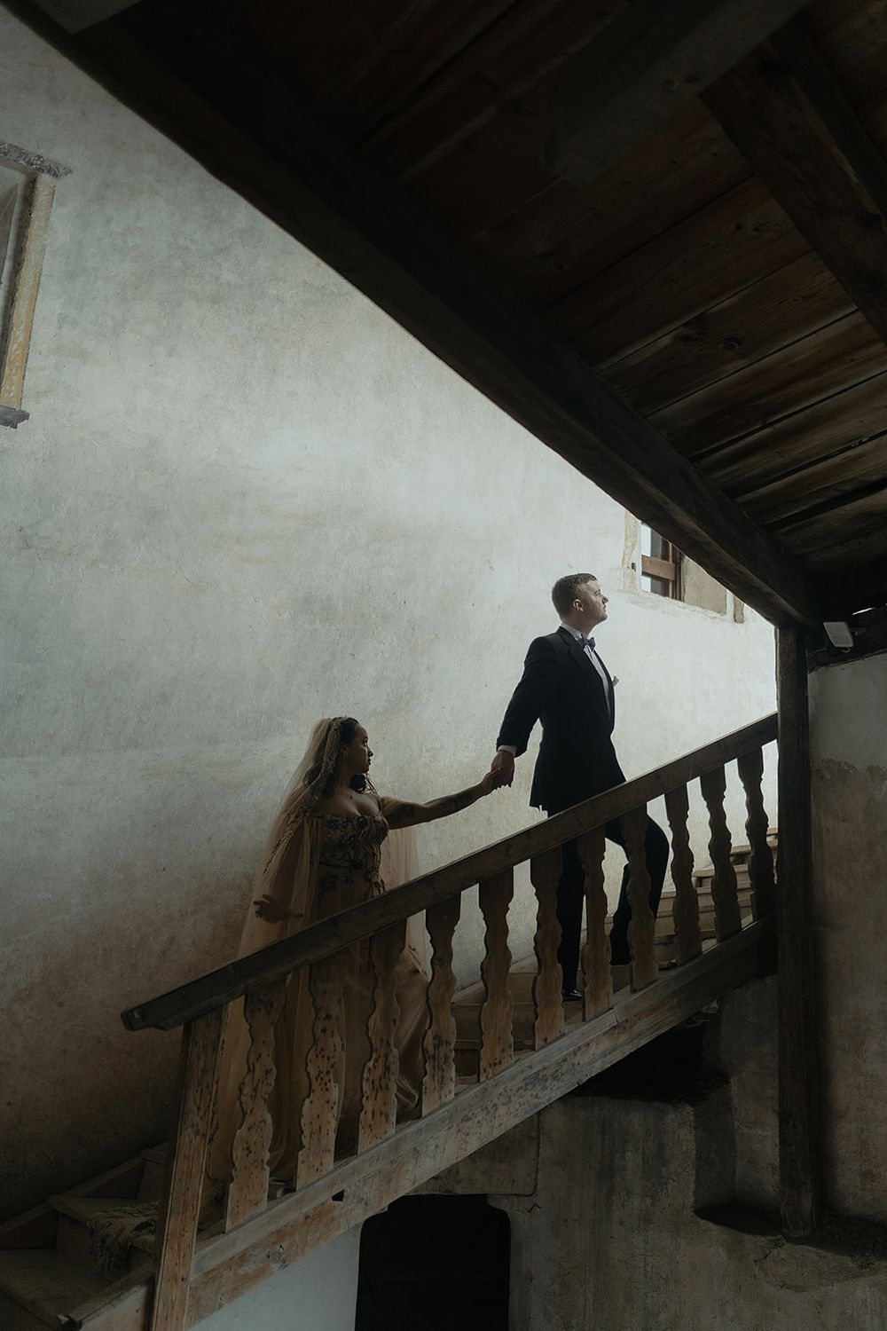 Bride and groom hold hands on rustic wooden staircase in elegant venue, captured in moody romantic wedding photography.