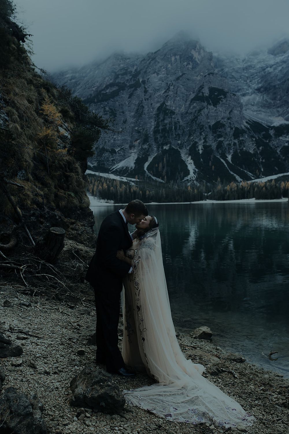 Couple sharing a romantic kiss in wedding attire by a misty alpine lake with dramatic mountain backdrop.