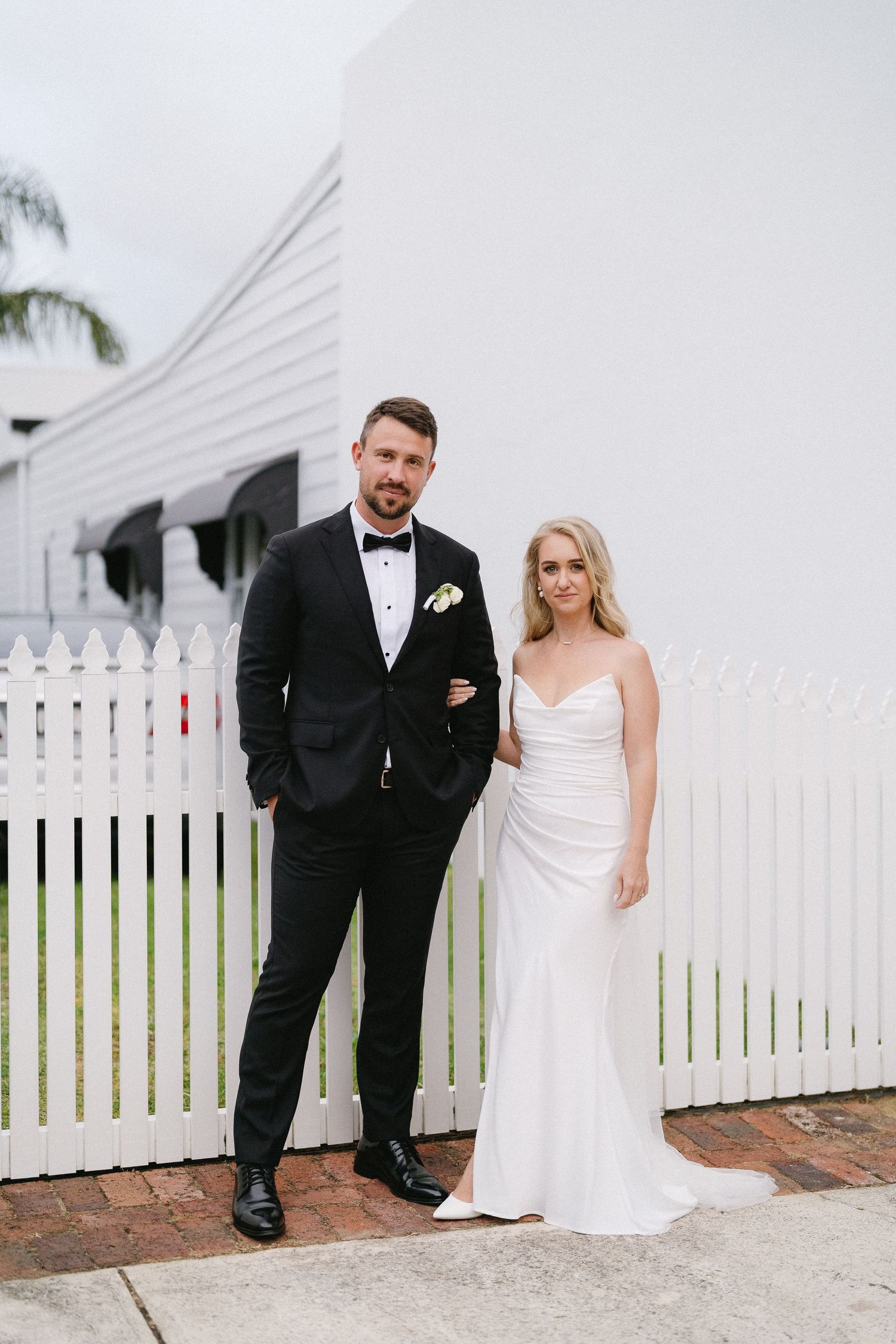 Bride in white strapless gown and groom in black tuxedo posing by white picket fence outside white building.