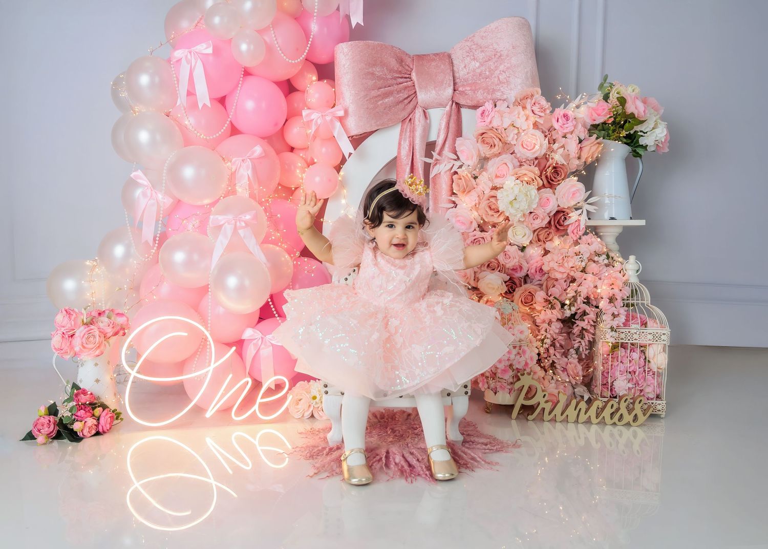 Baby girl in pink tutu dress sits among pink balloons and flowers at a Princess first birthday party.