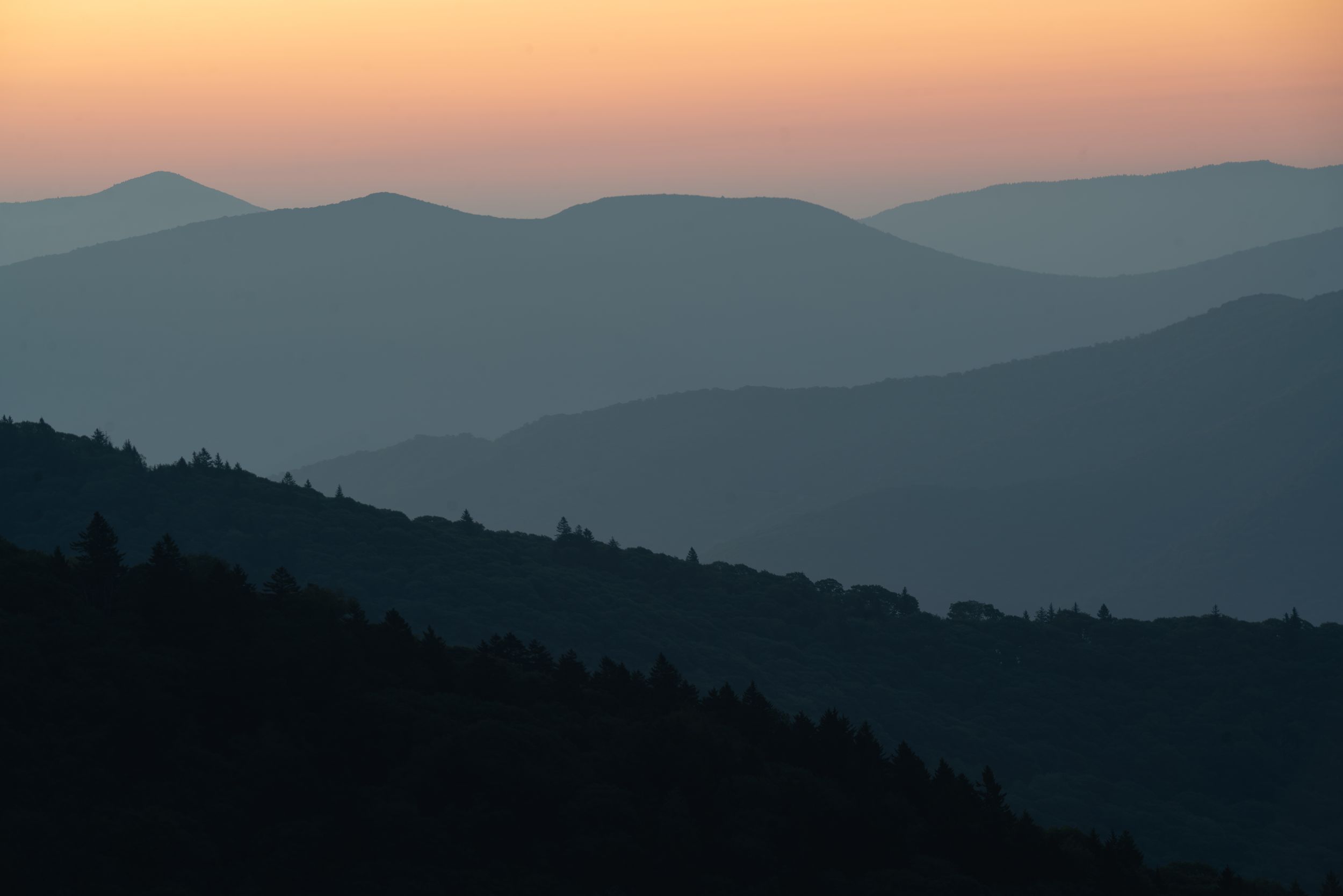 Layered blue mountain ridges fading into an orange and pink sunset horizon at dusk.
